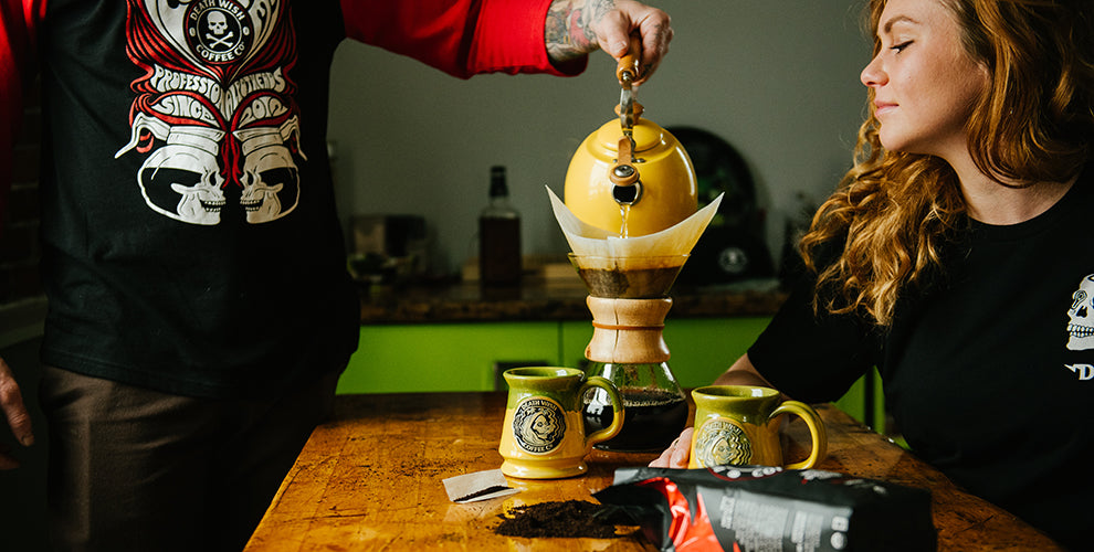 A man using the pour over method pouring hot water into a Chemex with two yellow mugs and a female waiting for the coffee.