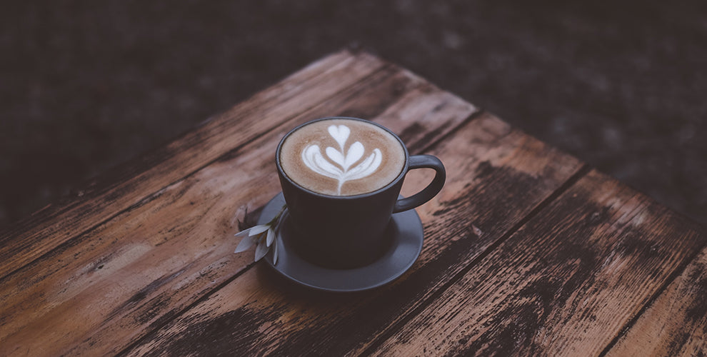 A latte in a black mug sitting on a wooden table that was recently stained using coffee.