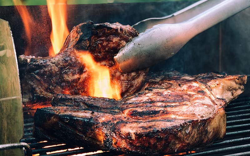 A steak on an open-flamed grill being flipped with metal tongs