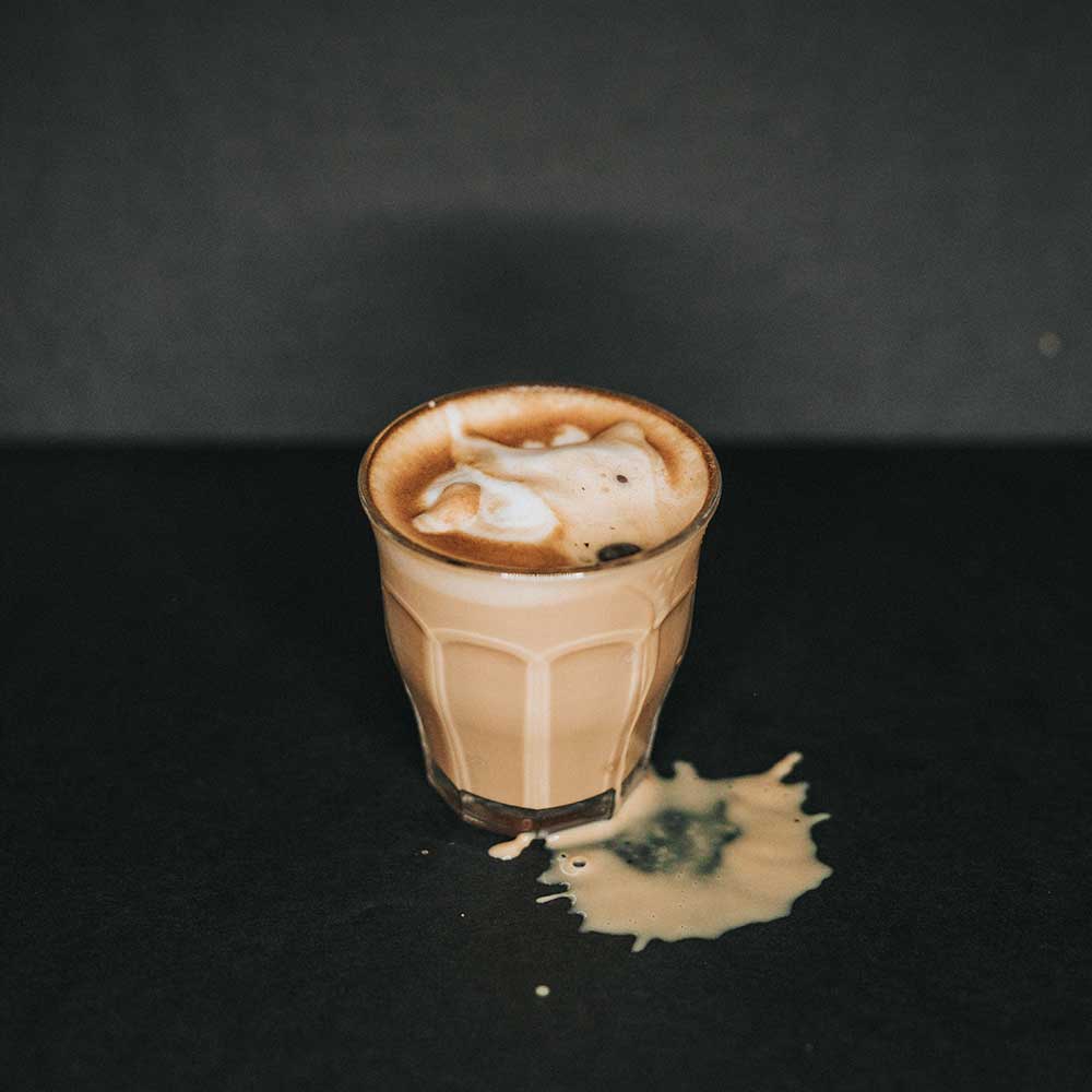 An image of keto coffee in a clear glass spilled on a back tabletop with a black background.