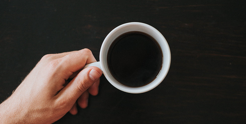 A hand holding a white mug on a black counter filled with black coffee.