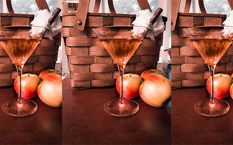 An image of three pumpking spiced martinis in a frosted martini glass with a cinnamon stick as a garnish. The backdrop is fall themed with a picnic basket and a few apples on a table. 