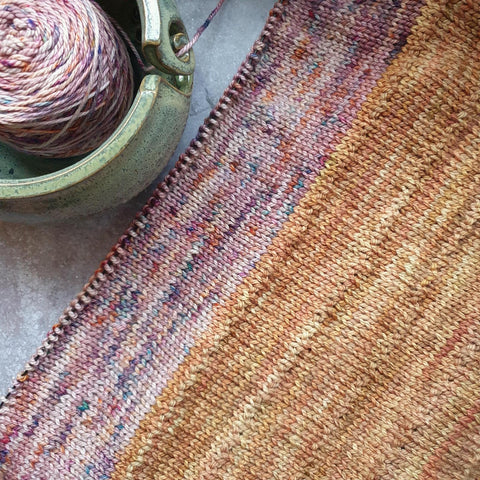The knitted fabric of a sweater on the needles, lying on the diagonal with a yarn bowl holding a cake of yarn in the corner. The fabric is made up of Gold and Rhino Tears.