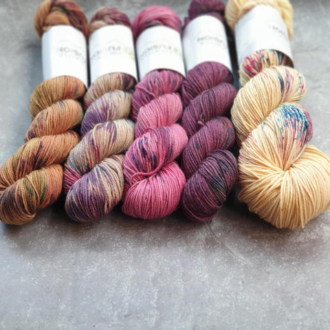 Four 50g skeins lined up next to a 100g skein. The colourways are variegated and from left to right they are Gold, Stone, Raspberry, Tulip and Honey Bloom.