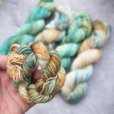 A variegated, cream, blue and rust, curled up mini skein in my hand, in the foreground, with for full size skeins in the background in similar colours