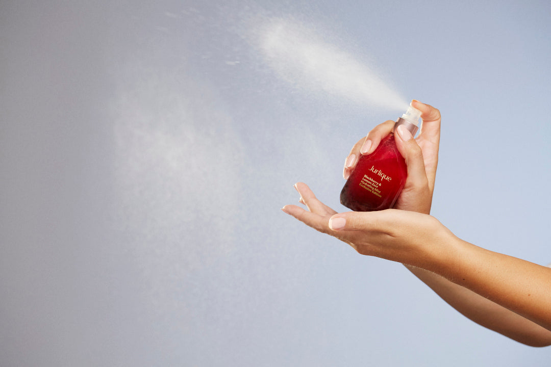 Woman’s hands holding red bottle of Jurlique Blackberry & Lemon Zest Hydrating Mist Exclusive Edition and spraying on blue background 