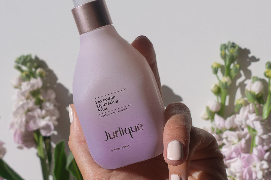 Woman’s hand holding purple bottle of Jurlique Lavender Hydrating Mist in front of a backdrop of flowers 