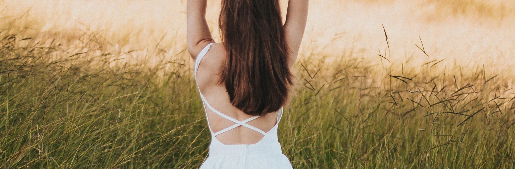 Bare back of woman with medium length brown hair in white dress on a grassy field.