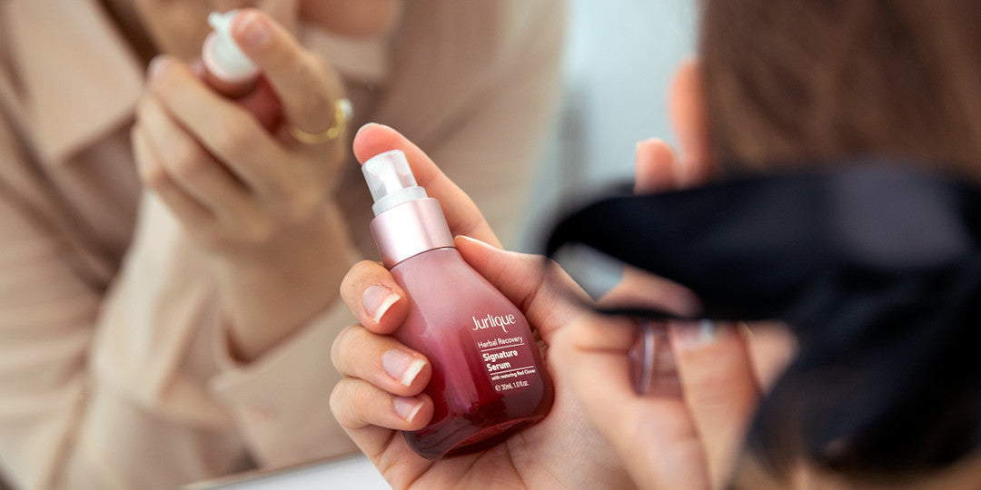 Woman looking in mirror and holding bottle of Jurlique Herbal Recovery Signature Serum