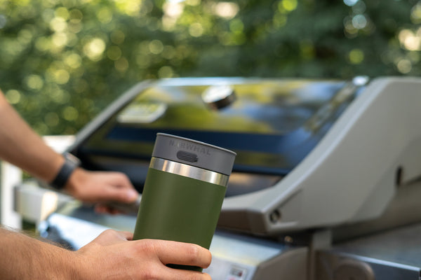 Man holds a green yeti tumbler with a NARWHAL bluetooth speaker tumbler lid in hand. A BBQ grill in the background against a Texas yard in summertime