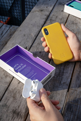 Yellow reboxed eco phone case and reboxed Apple AirPods Pro being held above the inside lid of a reboxed box on a wooden picnic bench