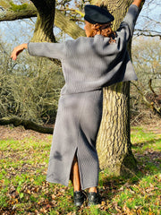 Outfit shot of person wearing matching grey skirt and jumper posing with arms above head facing away from camera