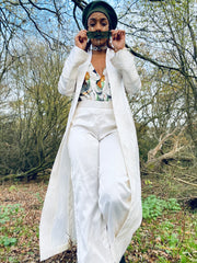 Outfit shot of woman wearing white trousers and white jacket with a green patterned top underneath facing forward with hands pulling down green tinted glasses and looking over the brim  posing