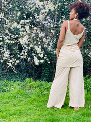 Outfit shot of person wearing beige strappy top and wide leg trousers facing the left side with tattoo on back and arms down 