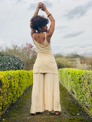 Outfit shot of person wearing beige strappy top and wide leg trousers facing backwards with both arms above head posing