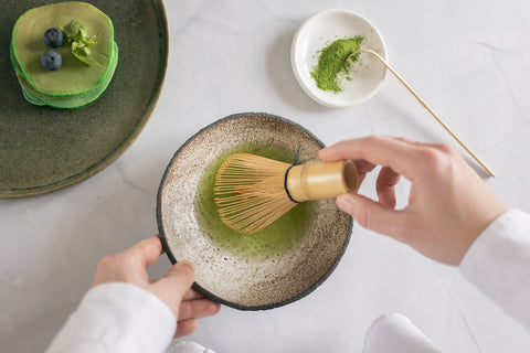 person whisking ceremonial matcha powder in bowl with bamboo whisk and matcha pancakes