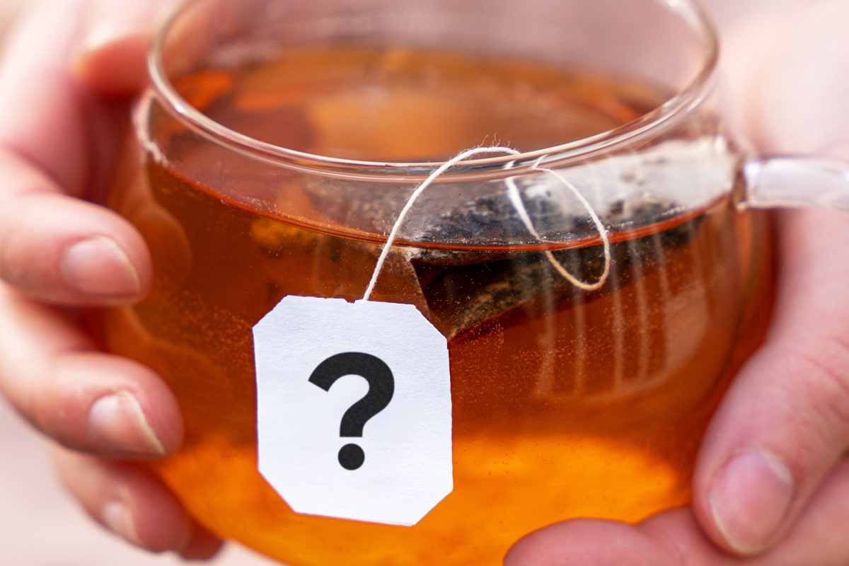 What to do with a teabag after drinking tea? Question illustrated by two hands cradling a glass mug of tea with the teabag still floating in the tea – there is a question mark on the tea-tag, which hangs over the side of the cup.