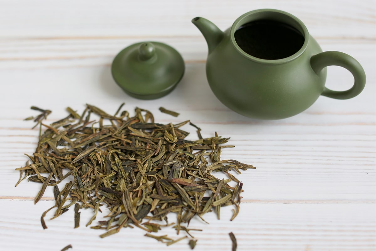 A green clay teapot with its lid off beside a small pile of Dragonfly Tea tea leaves on a white wooden surface.