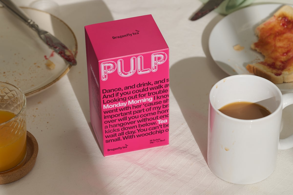 A pink box of Dragonfly Tea Pulp Monday Morning Tea on a breakfast table amongst a white mug of builder's tea, a glass of orange juice and a half eaten piece of jam-on-toast