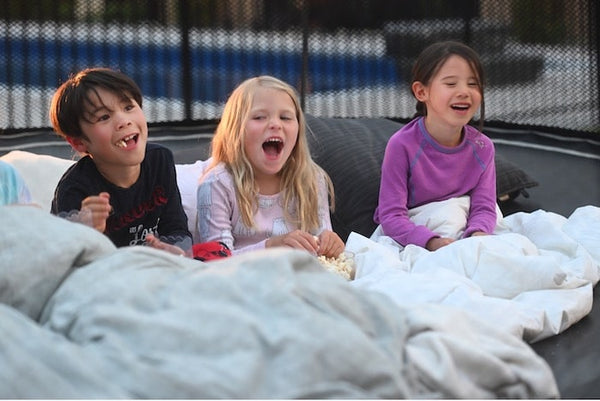 Three kids eating popcorn with blankets and pillows on a trampoline.