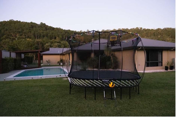 A Springfree Trampoline with a hoop, ball, ladder and lights.