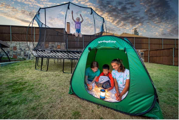 Three kids reading a book in a tent while another kid jumps on a Springfree Trampoline.