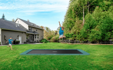 A kid jumping in mid-air on an inground trampoline while another kids run toward it.