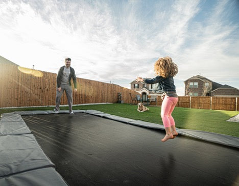A man and a little girl jumping on a rectangular inground trampoline.