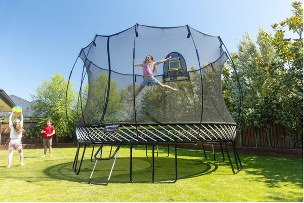 A girl jumping in mid-air on a Springfree Trampoline while two other kids throw a ball on the outside of it.