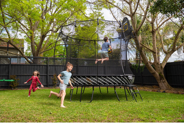 A boy dunking on a Springfree Trampoline Hoop while two other kids run around the Trampoline.