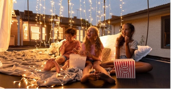 Three kids watching a movie on a trampoline with popcorn, blankets and lights.