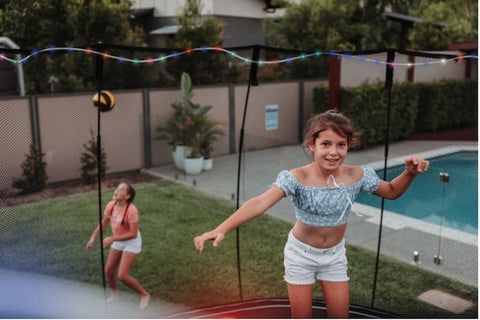 A girl jumping on a trampoline with lights while another girl catches a ball in the background.