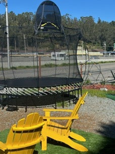 A Springfree Trampoline displayed outside next to a climbing dome and yellow chairs.