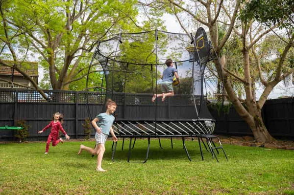 Two kids running around a Springfree Medium Round Trampoline while another kid jumps.