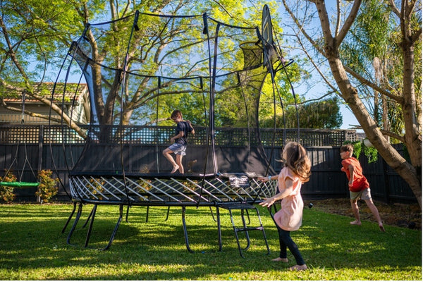 A child on a Springfree Trampoline while two other children play around it in a shaded backyard.