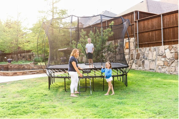 A boy jumping on a Springfree Trampoline while his mother and sister stand outside of it.
