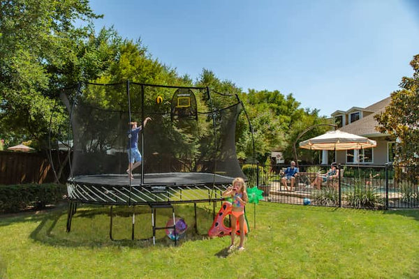 A boy shooting a basketball on a Springfree Trampoline while a girl plays with bubbles on the outside of it.