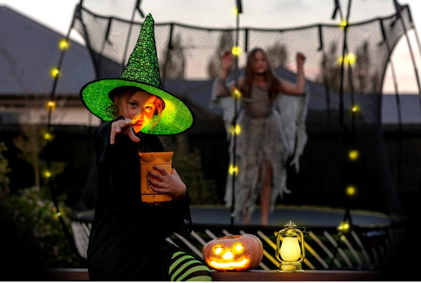 A girl dressed as a witch posing for the camera while another girl stands on a Springfree Trampoline in her costume.