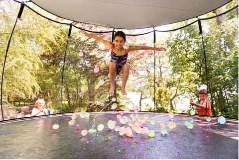 A little girl jumping on a trampoline with water balloons on the mat and a sunshade over the enclosure system.