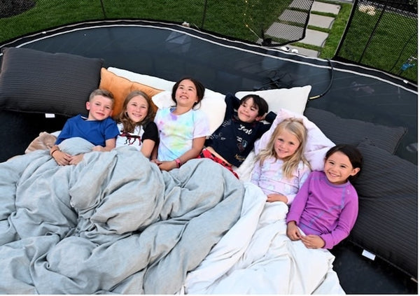 Multiple kids laying on a trampoline with pillows and blankets.
