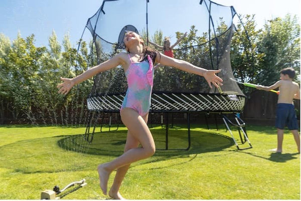 Kids playing by a sprinkler that's next to a Springfree Trampoline.