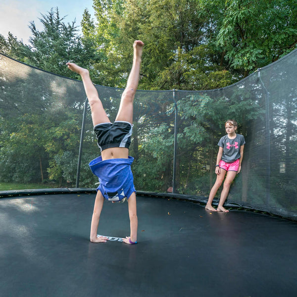 A girl doing a handstand on a trampoline while another girl leans against the net.