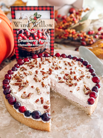 A cranberry pecan fluff cookie pie with a slice cut out on a marble turntable. A package of Wind & Willow cranberry pecan cheeseball mix. can be seen in the background with fall foliage.