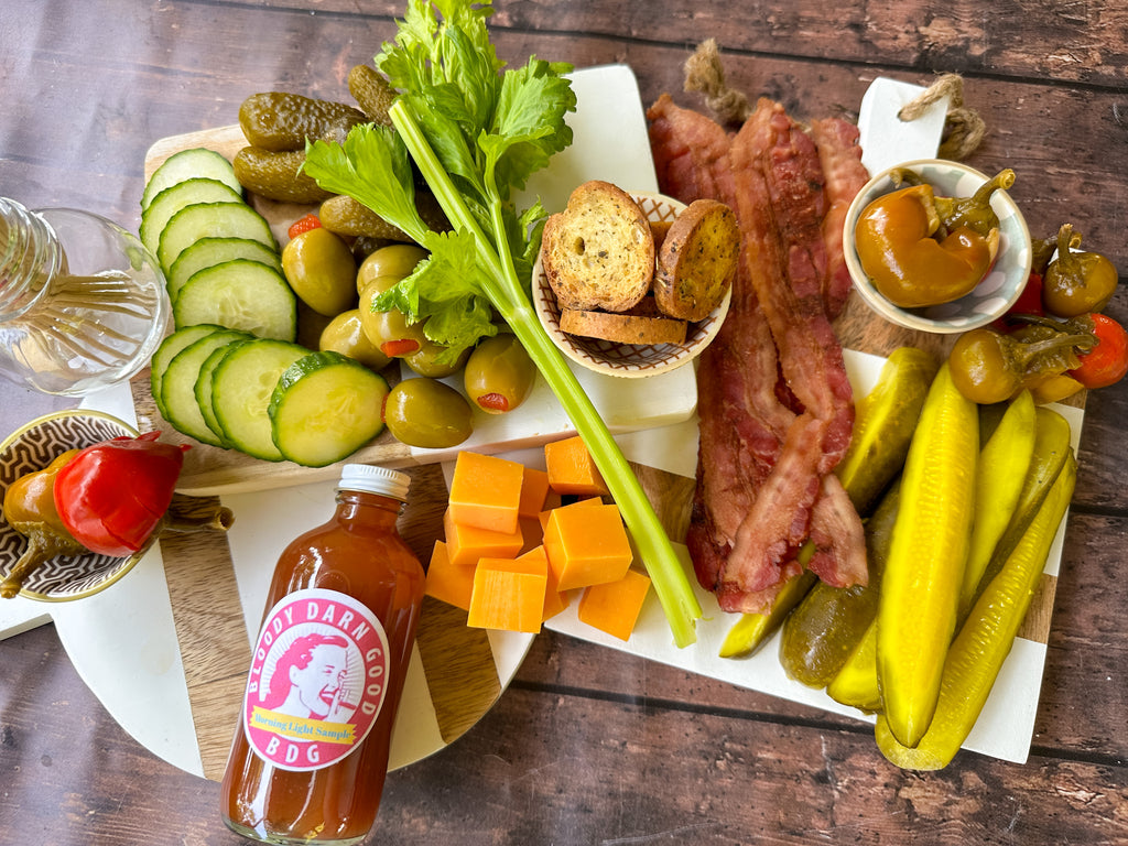 garnishes for a bloody mary cocktail including cucumber slices, peppers, pickles, celery, bacon, dill pickles, and cubes of cheese along with a sample bottle of Bloody Darn Good bloody mary mix.