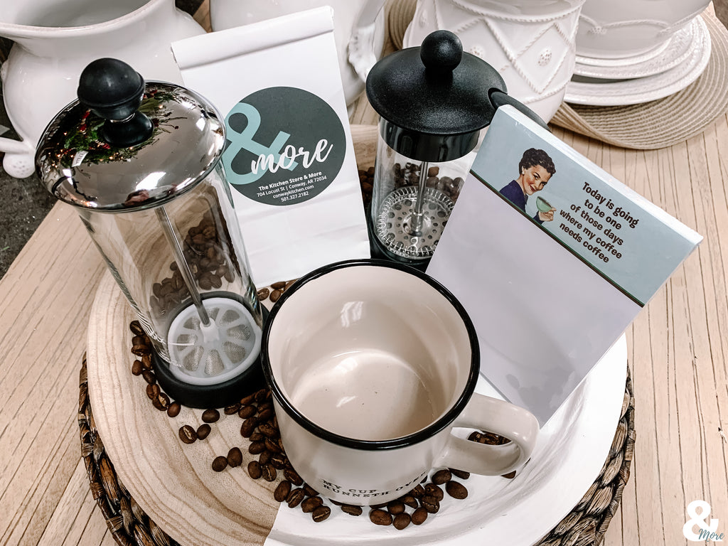 a round tray filled with coffee beans is loaded with a French press coffee maker and a glass milk frother. A bag of coffee with the & more label sits behind a coffee mug.