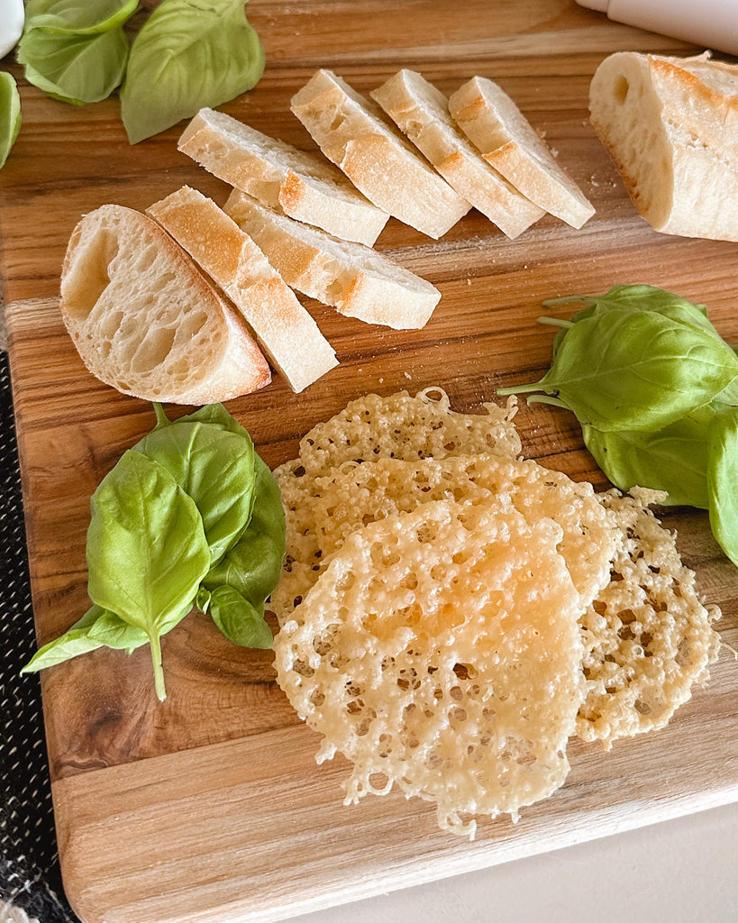 a stack of parmesan crisps on a cutting board along with sliced bread and basil leaves.