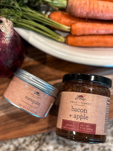 jars of finch + fennel mesquite seasoning and bacon + apple jam on a cutting board with carrots and an onion