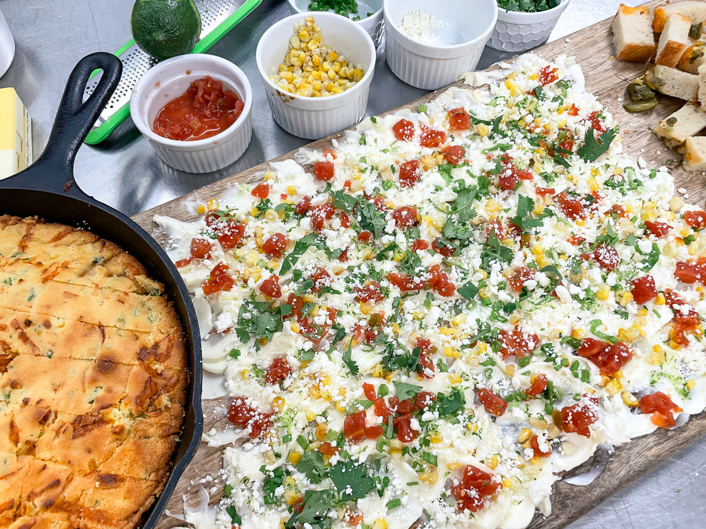 butter board with butter spread topped with corn, tomatoes, cilantro, green onion, lime zest and cotija cheese next to a cast iron skillet of cornbread