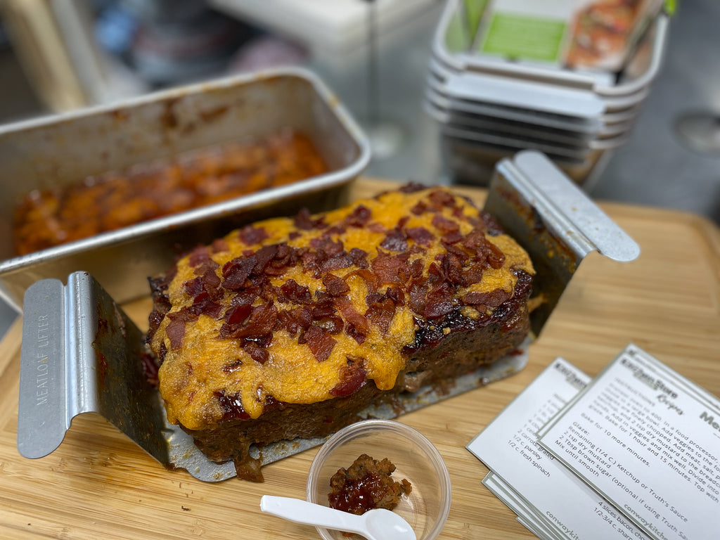 a bacon and cheese topped meatloaf sits in a NordicWare insert on a cutting board with recipe cards and a sample bite of meatloaf. 