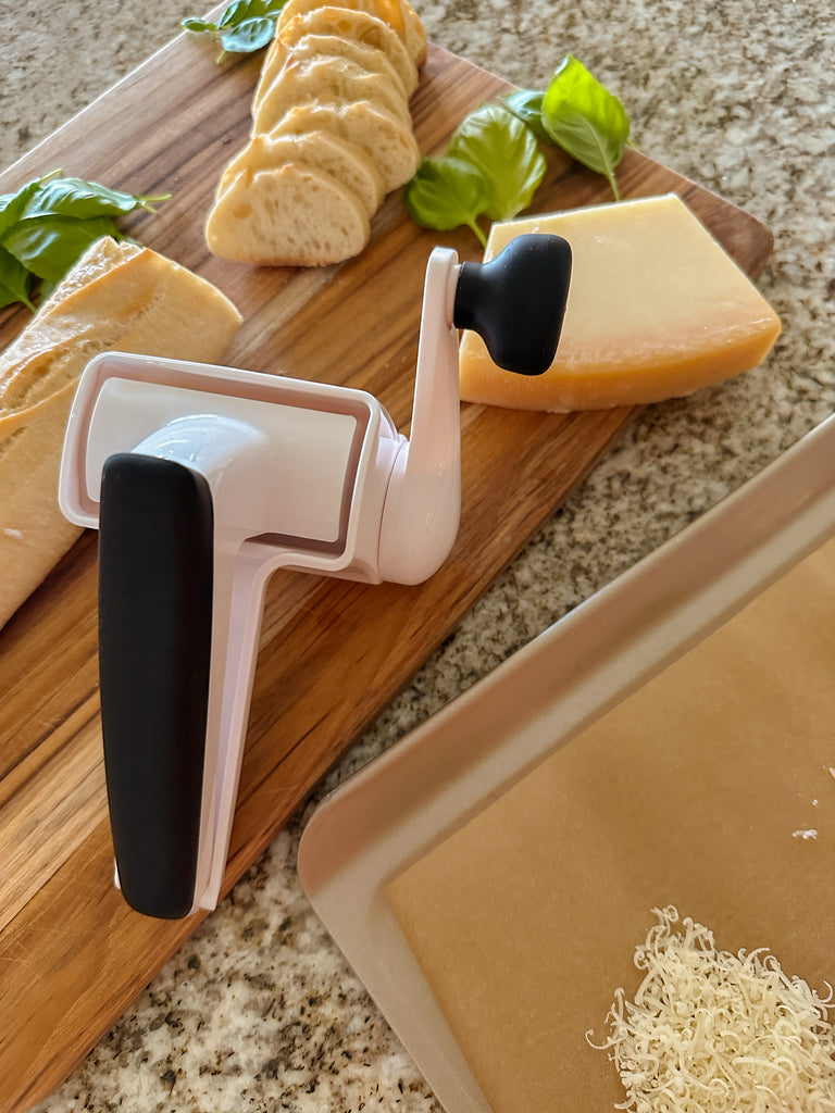 The OXO rotary grinder on a cutting board with parmesan cheese, slices of bread and basil leaves.